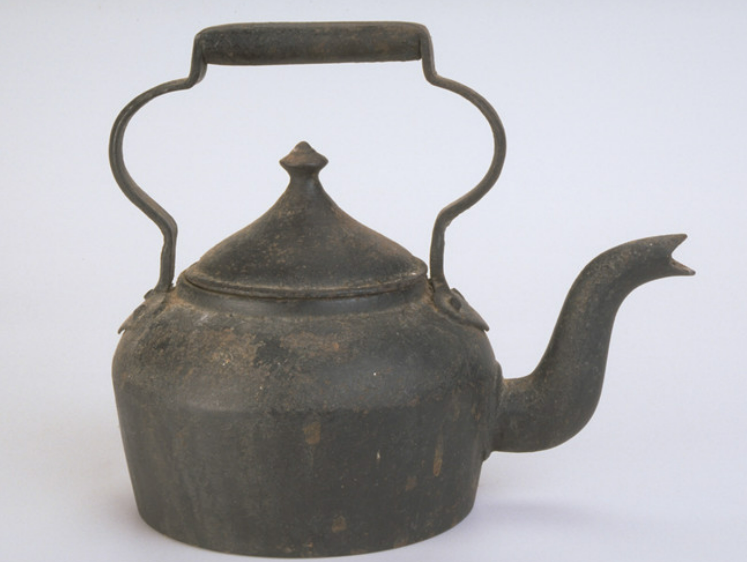 Photograph of old iron kettle with a curved pout and handles. 